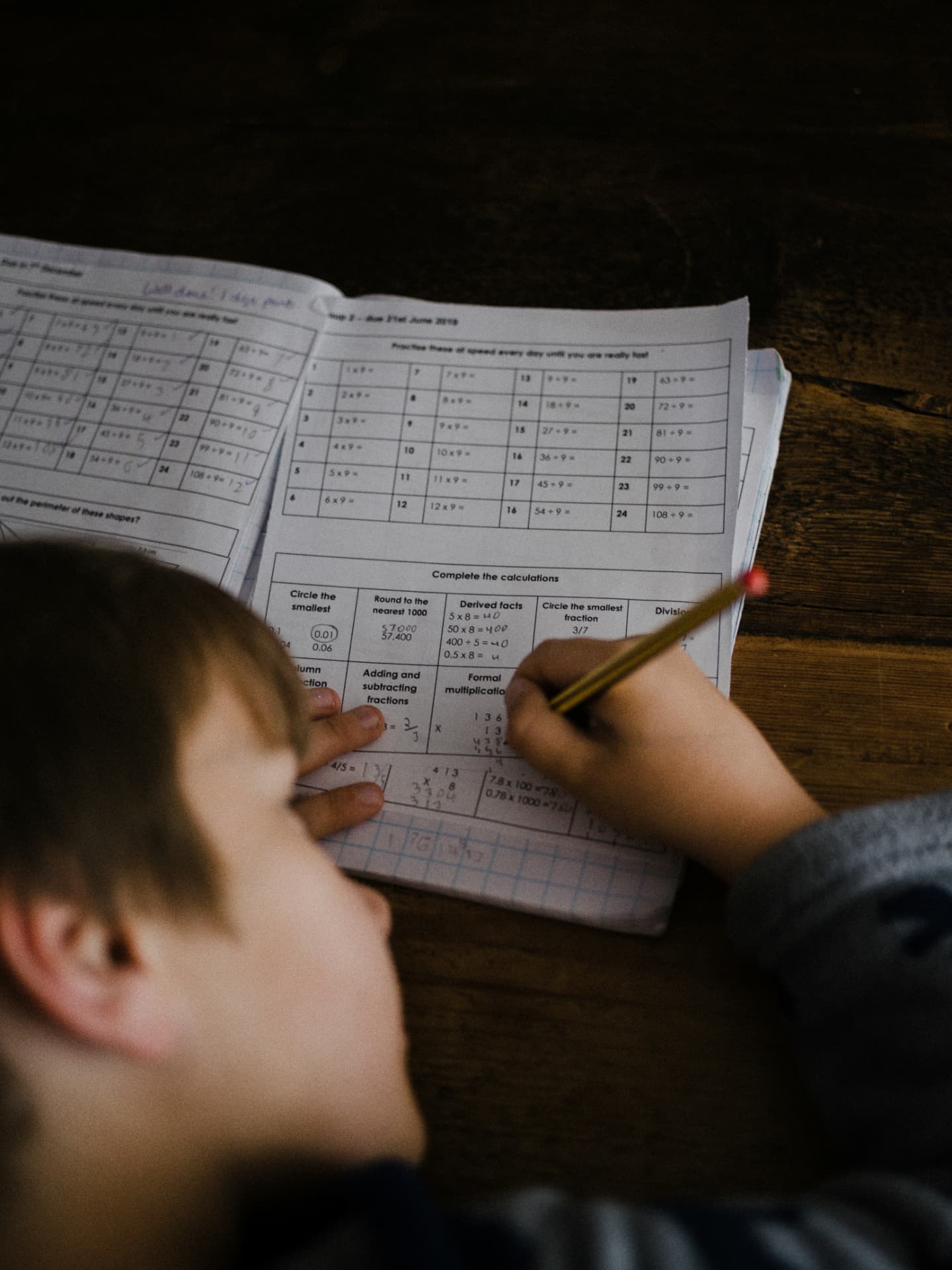A young student working through a maths workbook at a dark wooden desk, pencil in hand, with a handwritten Well done note at the top of the page
